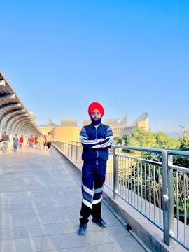 A person wearing a red turban and a blue tracksuit stands on a walkway bordered by metallic railings. The clear blue sky stretches above, while an architectural building with unique shapes is visible in the background. Several people are present further down the walkway, some dressed in colorful attire, walking in both directions.
