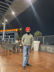 A man wearing a red turban and patterned long-sleeve shirt stands on a tiled terrace at night. Behind him are patio chairs and tables, large potted plants, and a building with 'Taj' signage. The area is well-lit with modern overhead lights.