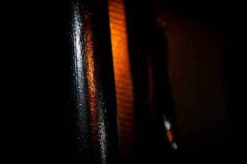 The image features close-up sections of black metallic or reflective surfaces illuminated by a warm, amber light from the right side. The lighting creates a contrasting effect between the dark areas and the orange glow, emphasizing texture and sheen.