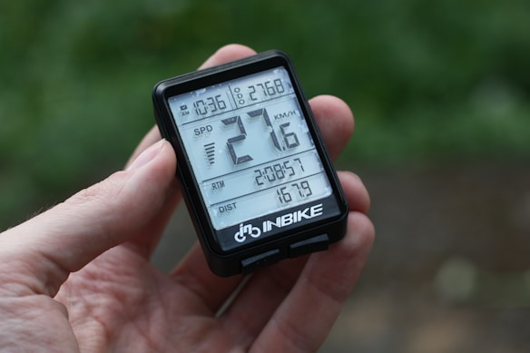 A hand is holding a digital cycling computer with a display showing time, speed in kilometers per hour, ride time, and distance covered. The device has a black casing and is branded with 'INBIKE' at the bottom.