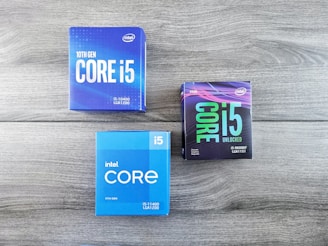 The image features three Intel Core i5 processor boxes arranged on a wooden surface. The boxes display details about the generation and type of processors, including the 10th Gen i5-10400, 11th Gen i5-11400, and 9th Gen i5-9600KF. Each box has distinct color schemes and branding.