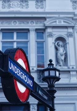 A London Underground sign with a red and blue color scheme is prominently displayed in the foreground. The sign is attached to a black metal lamp post. In the background, there is a historic-looking building with ornate architectural details and sculptures.
