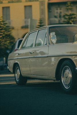 A vintage car is parked on a street with a close-up view of its side profile, showcasing the smooth lines and classic design. The background includes a building with large windows and small trees, indicating an urban setting. The lighting gives a warm, nostalgic atmosphere.