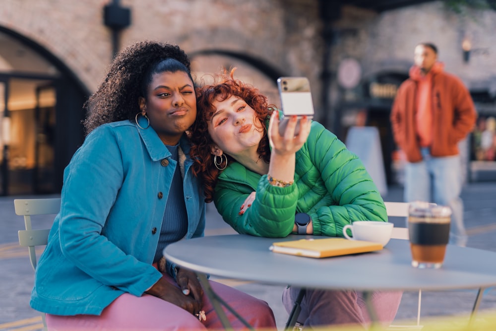 Una persona que se toma una selfie con otra mujer sentada en una mesa