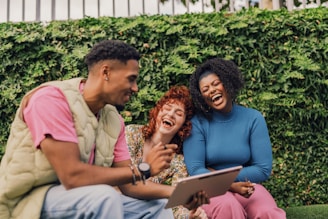 Three people sitting outdoors, engaged in a cheerful conversation. They are laughing together, creating a light-hearted and joyful atmosphere. The background is filled with lush green foliage, which adds a natural and vibrant setting to their interaction. One person is holding a tablet, and everyone appears relaxed and happy.