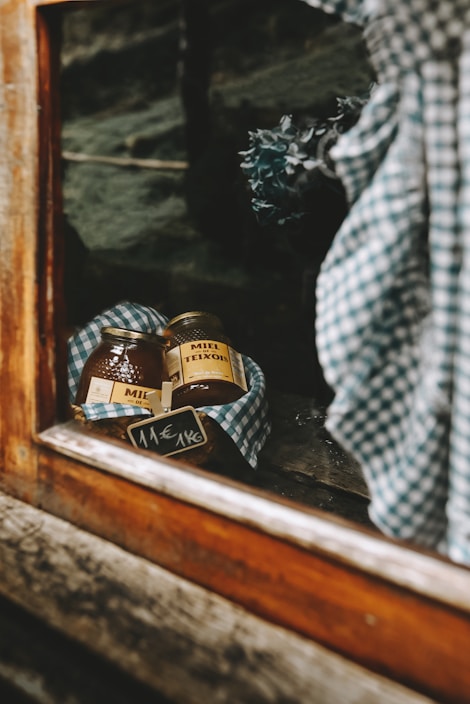 Two jars of honey wrapped in a checkered cloth are displayed on a shelf behind a window. A small price tag indicating 11€ per kg is visible. The setting appears rustic, with wooden elements framing the scene, and a bouquet of dried flowers slightly out of focus in the background.