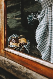 Two jars of honey wrapped in a checkered cloth are displayed on a shelf behind a window. A small price tag indicating 11&euro; per kg is visible. The setting appears rustic, with wooden elements framing the scene, and a bouquet of dried flowers slightly out of focus in the background.