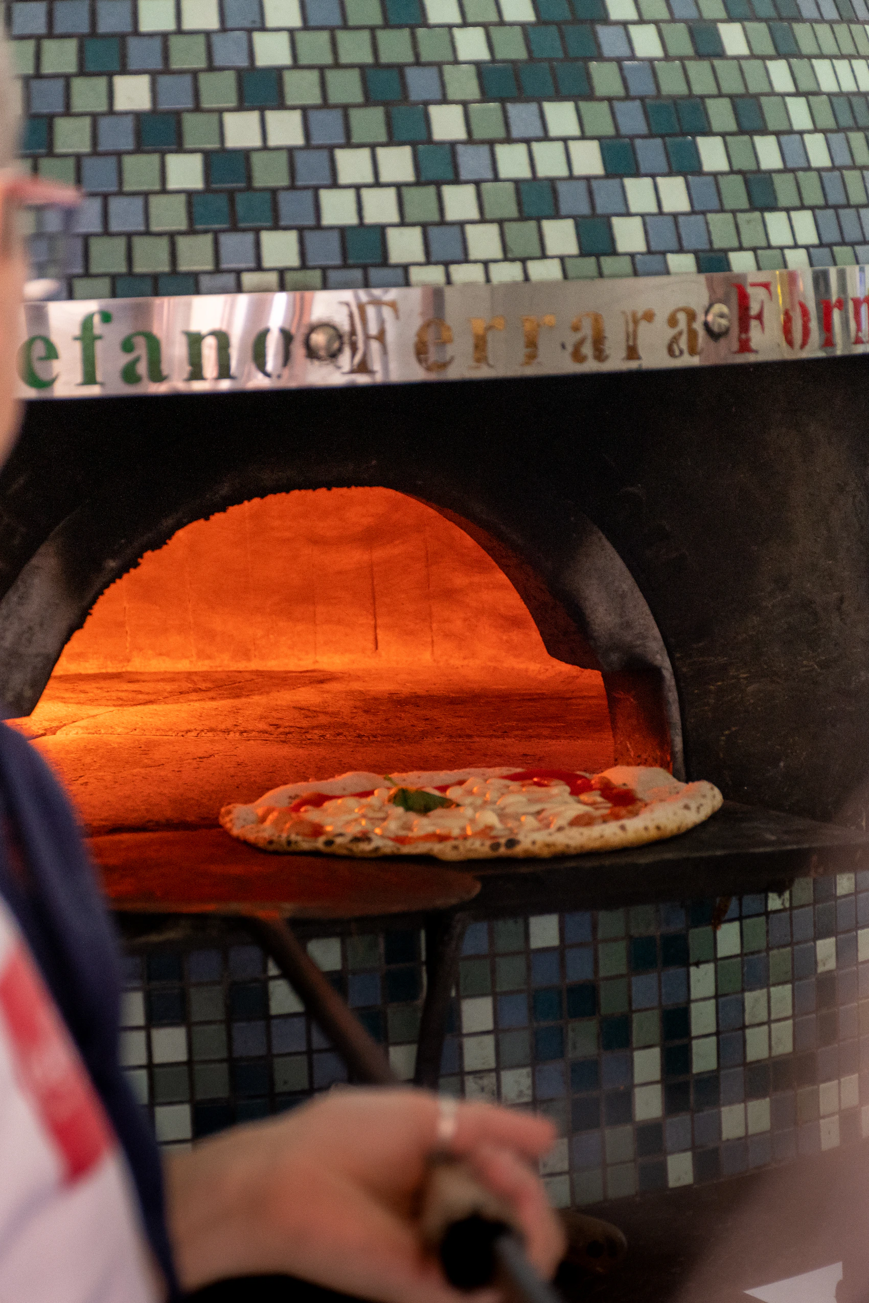 A pizza maker sliding a Neapolitan pizza into a glowing tiled wood-fired oven