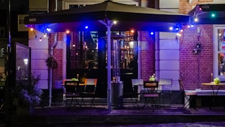 An outdoor cafe scene with several tables and chairs arranged under an awning. The cafe is adorned with colorful string lights creating a cozy ambiance. The brick walls and glass doors in the background suggest a quaint urban setting. Small plants or flowers decorate the tables, adding a touch of greenery to the scene.