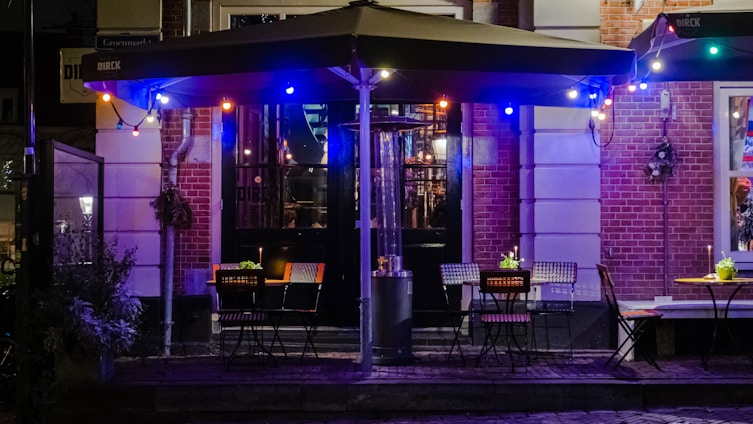 An outdoor cafe scene with several tables and chairs arranged under an awning. The cafe is adorned with colorful string lights creating a cozy ambiance. The brick walls and glass doors in the background suggest a quaint urban setting. Small plants or flowers decorate the tables, adding a touch of greenery to the scene.