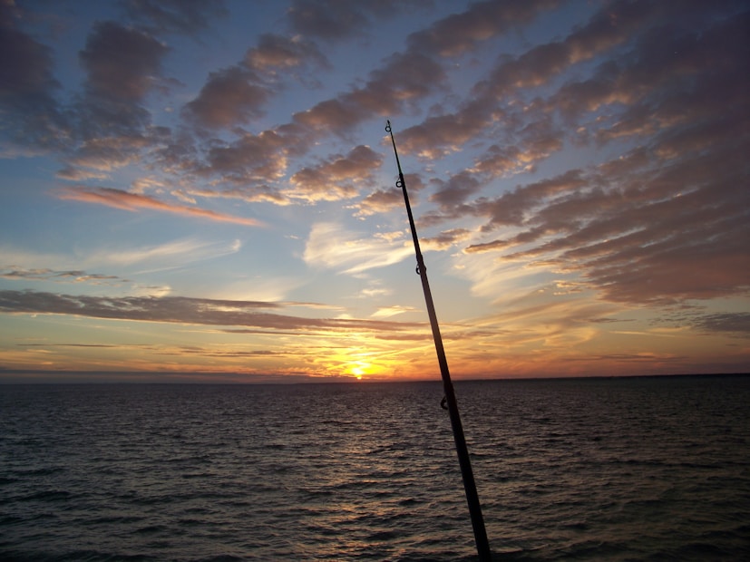 A fishing rod is silhouetted against a dramatic sunset over the ocean, with numerous clouds partially covering the vibrant sky, creating a picturesque scene.