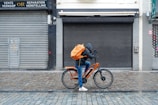 A cyclist rides along a cobblestone street, wearing a hooded jacket and carrying a large orange backpack. The bicycle is also orange, complementing the backpack. The scene is set against a backdrop of closed shops with gray metal shutters and a tiled sidewalk.