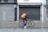 A cyclist rides along a cobblestone street, wearing a hooded jacket and carrying a large orange backpack. The bicycle is also orange, complementing the backpack. The scene is set against a backdrop of closed shops with gray metal shutters and a tiled sidewalk.