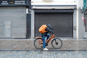 A cyclist rides along a cobblestone street, wearing a hooded jacket and carrying a large orange backpack. The bicycle is also orange, complementing the backpack. The scene is set against a backdrop of closed shops with gray metal shutters and a tiled sidewalk.