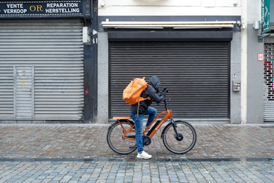 A cyclist rides along a cobblestone street, wearing a hooded jacket and carrying a large orange backpack. The bicycle is also orange, complementing the backpack. The scene is set against a backdrop of closed shops with gray metal shutters and a tiled sidewalk.