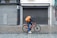 A cyclist rides along a cobblestone street, wearing a hooded jacket and carrying a large orange backpack. The bicycle is also orange, complementing the backpack. The scene is set against a backdrop of closed shops with gray metal shutters and a tiled sidewalk.