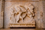 A detailed stone carving depicting a mythological scene with figures holding weapons, possibly from an ancient epic or storytelling. The characters exhibit dynamic poses, suggesting action and movement.