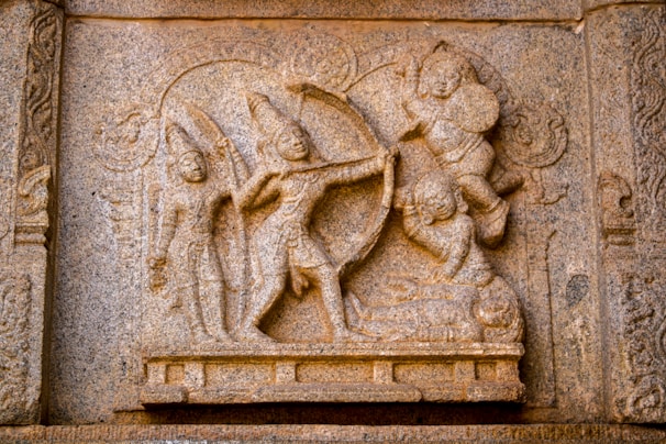 A detailed stone carving depicting a mythological scene with figures holding weapons, possibly from an ancient epic or storytelling. The characters exhibit dynamic poses, suggesting action and movement.