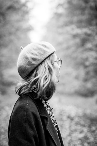 A black and white image featuring a person in a side profile. The individual is wearing a beret and glasses, with shoulder-length hair. Trees and foliage create a blurred background, suggesting an outdoor setting.