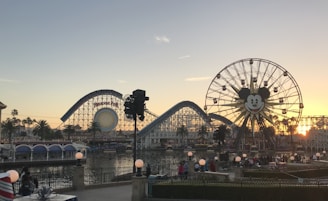 A coastal amusement park scene features a large Ferris wheel with a smiling face in the center. Roller coasters and other amusement rides are visible, along with palm trees and a body of water reflecting the sunset. Visitors can be seen walking and enjoying the surroundings.