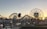 A coastal amusement park scene features a large Ferris wheel with a smiling face in the center. Roller coasters and other amusement rides are visible, along with palm trees and a body of water reflecting the sunset. Visitors can be seen walking and enjoying the surroundings.
