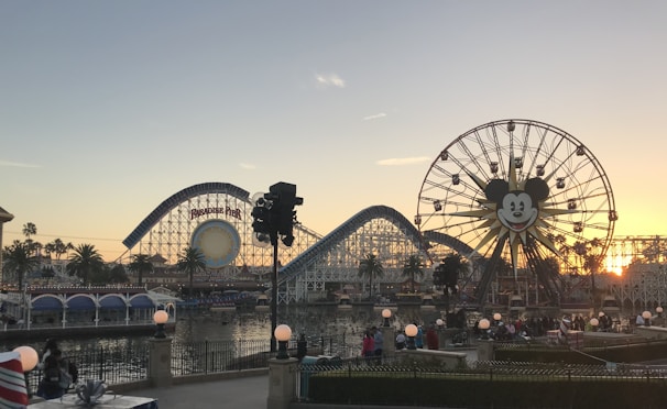 A coastal amusement park scene features a large Ferris wheel with a smiling face in the center. Roller coasters and other amusement rides are visible, along with palm trees and a body of water reflecting the sunset. Visitors can be seen walking and enjoying the surroundings.