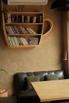A cozy reading nook is featured with a unique bookshelf shaped like a teacup, filled with various books. Below it, there's a dark leather couch adorned with green cushions. A wooden table is placed in front of the couch, and a potted plant is visible on the left. The walls and furniture create a warm, inviting atmosphere.