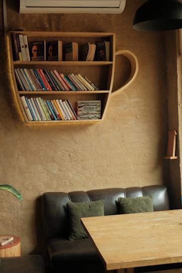 A cozy reading nook is featured with a unique bookshelf shaped like a teacup, filled with various books. Below it, there's a dark leather couch adorned with green cushions. A wooden table is placed in front of the couch, and a potted plant is visible on the left. The walls and furniture create a warm, inviting atmosphere.