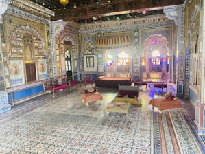 An ornately decorated room showcases intricate wall patterns, detailed carvings, and vibrant stained glass windows. The room is furnished with traditional wooden furniture, including chairs and low tables covered with cushions in warm colors. The décor reflects a rich historical and cultural ambiance.