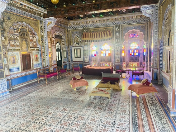 An ornately decorated room showcases intricate wall patterns, detailed carvings, and vibrant stained glass windows. The room is furnished with traditional wooden furniture, including chairs and low tables covered with cushions in warm colors. The décor reflects a rich historical and cultural ambiance.