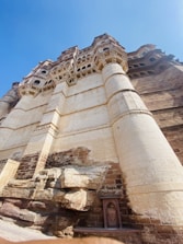 A majestic fortress structure is shown with intricate carvings and large cylindrical columns. The towering architecture is constructed from a beige and brown stone, featuring a mix of smooth and textured surfaces. Underneath, natural rock formations add to the grandeur.