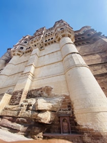 A majestic fortress structure is shown with intricate carvings and large cylindrical columns. The towering architecture is constructed from a beige and brown stone, featuring a mix of smooth and textured surfaces. Underneath, natural rock formations add to the grandeur.