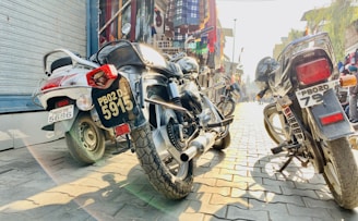 Three motorcycles are parked on a cobblestone street, with a variety of colorful fabrics and garments displayed in the background on hanging racks. Sunlight casts distinct shadows on the pavement, conveying a bright, bustling atmosphere.