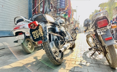 Three motorcycles are parked on a cobblestone street, with a variety of colorful fabrics and garments displayed in the background on hanging racks. Sunlight casts distinct shadows on the pavement, conveying a bright, bustling atmosphere.