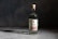 A bottle of Wild Common mezcal is placed on a smooth, textured surface with a dark, moody background. The bottle is clear, showcasing the transparent liquid inside, and features a label with the brand name and product details. The lighting highlights the contours of the bottle, casting a subtle shadow.