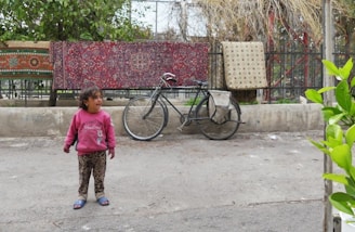 A child wearing a pink sweatshirt and leopard print pants stands on a pavement. Behind the child, two colorful rugs are hanging on a metal fence, and there is a bicycle leaning against the fence. The surroundings include some greenery to the right and trees beyond the fence.