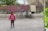 A child wearing a pink sweatshirt and leopard print pants stands on a pavement. Behind the child, two colorful rugs are hanging on a metal fence, and there is a bicycle leaning against the fence. The surroundings include some greenery to the right and trees beyond the fence.