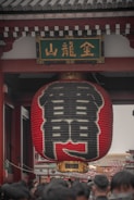 A large red Japanese lantern with black kanji characters hangs prominently under a traditional temple gate. The architectural elements suggest a significant cultural or historic site. A crowd of people is gathered beneath, possibly visitors or tourists.