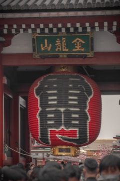 A large red Japanese lantern with black kanji characters hangs prominently under a traditional temple gate. The architectural elements suggest a significant cultural or historic site. A crowd of people is gathered beneath, possibly visitors or tourists.