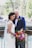 A couple stands together in wedding attire, with the bride wearing a white lace gown and veil, holding a vibrant bouquet of colorful flowers. The groom is dressed in a navy blue suit with a white shirt and orange bow tie. They pose in an outdoor setting with green trees and a bridge in the background.