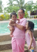 An adult wearing glasses and pink clothing holds a smiling baby while another young child, also in pink, stands beside them. They are near a swimming pool with lush greenery in the background.