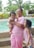 An adult wearing glasses and pink clothing holds a smiling baby while another young child, also in pink, stands beside them. They are near a swimming pool with lush greenery in the background.