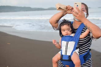 A woman wearing a face mask and a striped shirt holds a phone in one hand while adjusting her hair with the other. She carries a baby in a blue carrier on her chest. The setting is a beach with waves in the background.