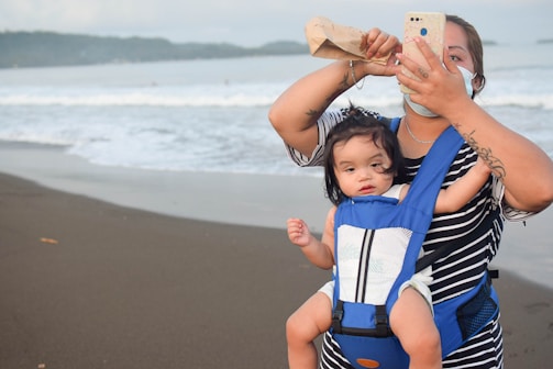 A woman wearing a face mask and a striped shirt holds a phone in one hand while adjusting her hair with the other. She carries a baby in a blue carrier on her chest. The setting is a beach with waves in the background.