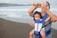 A woman wearing a face mask and a striped shirt holds a phone in one hand while adjusting her hair with the other. She carries a baby in a blue carrier on her chest. The setting is a beach with waves in the background.