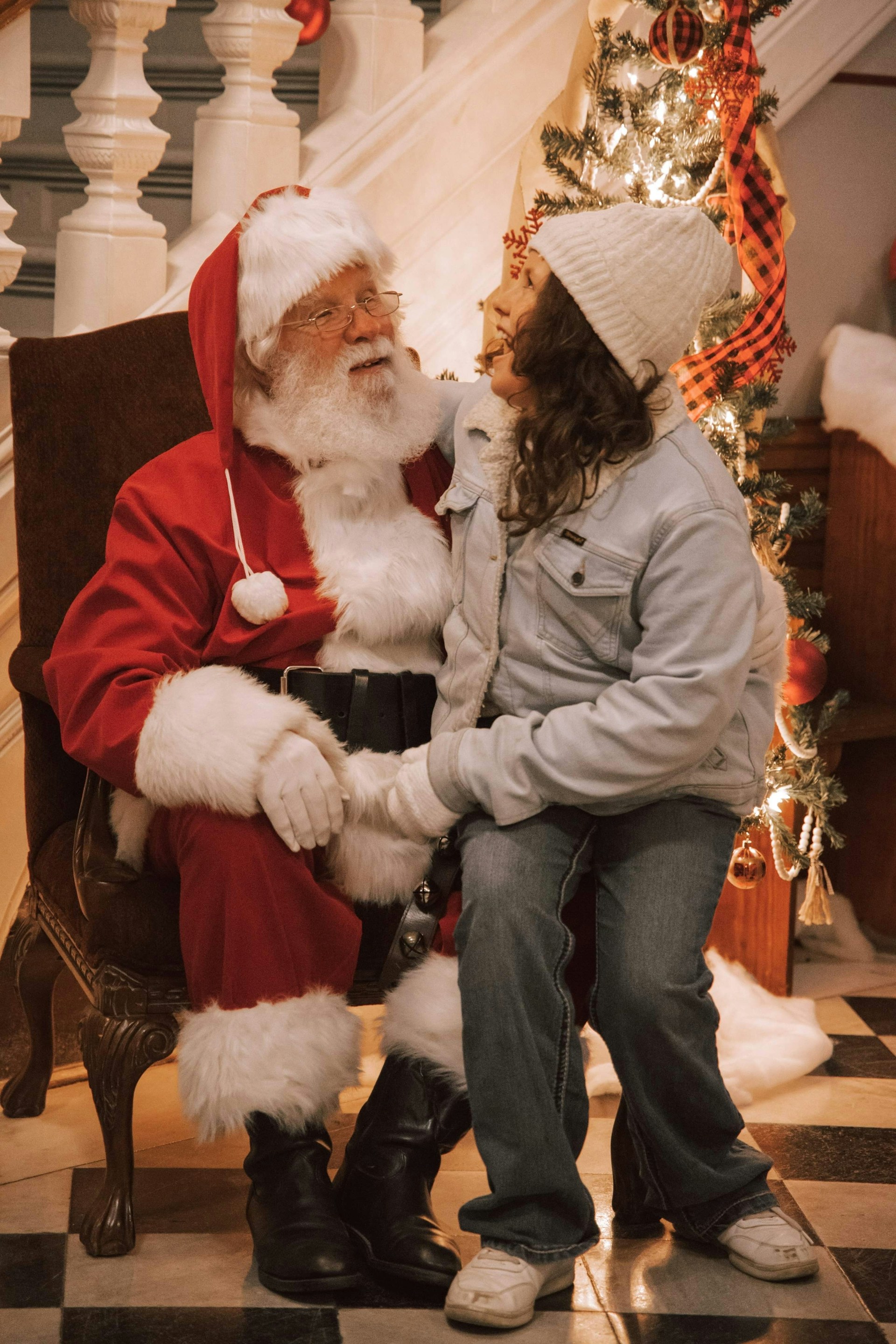 A person dressed as Santa Claus is sitting on a chair with a young person on their lap. They are in a festive setting with a decorated Christmas tree, which is illuminated with lights. The atmosphere is warm and cozy, indicative of the holiday season.