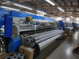 An industrial setting with multiple large textile weaving machines placed in a row, surrounded by packages and various materials. The machines are primarily blue and white, featuring several moving components and long rollers. The area is well-lit by overhead lights and is spacious with a concrete floor.