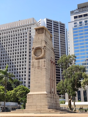 A tall stone cenotaph with inscriptions stands prominently in front of modern high-rise buildings. Trees surround the structure, and the sky is clear and sunny.