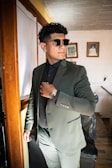 A person wearing a dark suit and sunglasses stands confidently in a well-lit room. The individual is adjusting their suit collar while looking to the side. The room has framed pictures on the wall and wooden furniture visible.