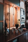 A wooden shelf displays hanging macramé art and a vase with dried branches. Below them, a collection of ceramic cups, a plant in a terracotta pot, bottles, and framed black and white photographs provide a cozy, rustic vibe.